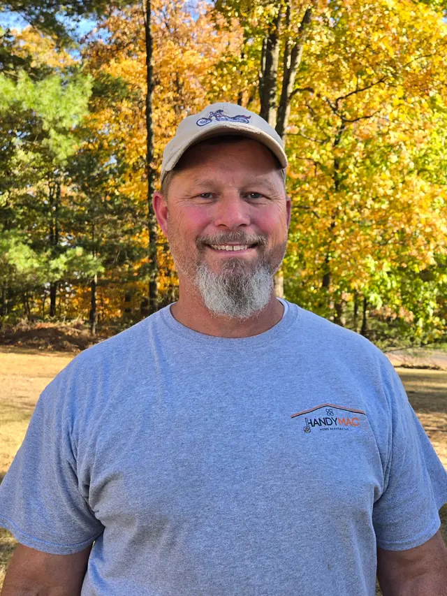 Joel MacLeod in a cap and a HandyMac T-shirt in front of autumn trees