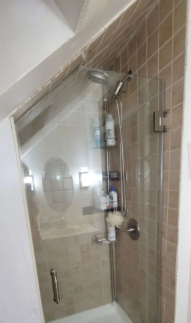 A frameless glass shower door with a steel hinge in a tiled shower under a sloped ceiling